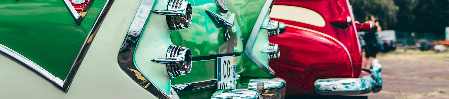 The image shows the rear ends of two vintage cars, one green and one red, parked side by side on a sunny day.