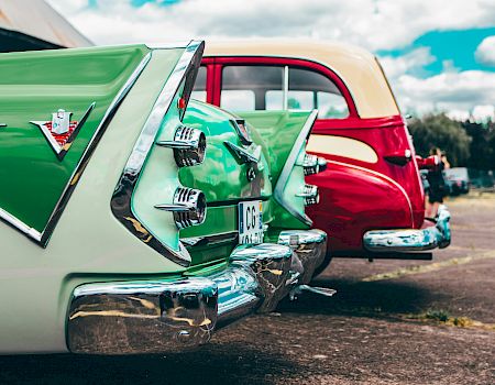 The image shows the rear ends of two vintage cars, one green and one red, parked side by side on a sunny day.