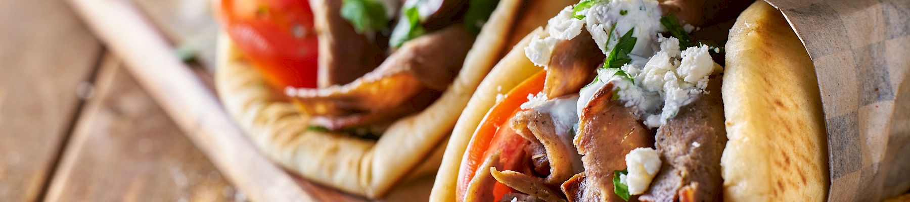 The image shows two gyro sandwiches filled with meat, vegetables, and sauce, wrapped in paper, and served with French fries on a wooden surface.