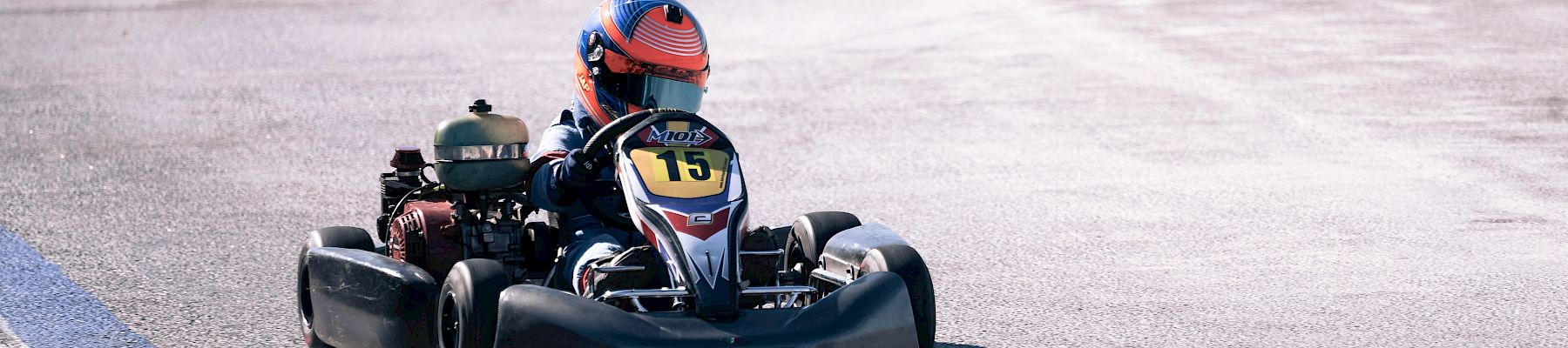 A person wearing a helmet drives a go-kart labeled with the number 15 on a paved track with blue and white markings.