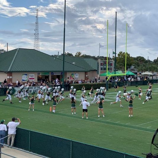 A high school football field with cheerleaders performing, players in formation, and spectators in bleachers under a partly cloudy sky.
