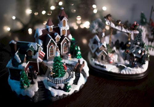 A miniature Christmas village with snowy decorations, trees, and a church, set against twinkling lights in the background.