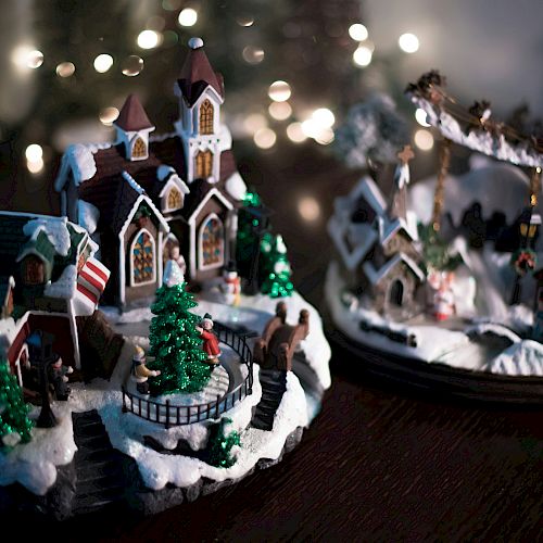 A miniature Christmas village with snowy decorations, trees, and a church, set against twinkling lights in the background.