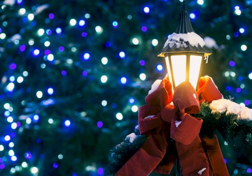 A snow-covered lamp post adorned with a red bow and wreath, set against a backdrop of blue holiday lights on a tree.