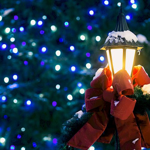 A snow-covered lamp post adorned with a red bow and wreath, set against a backdrop of blue holiday lights on a tree.