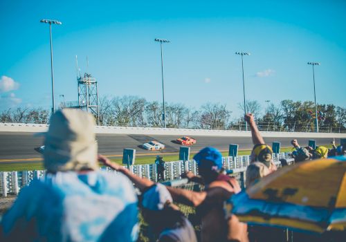 Spectators cheer at a car race on a sunny day, with three race cars speeding on the track in the background.