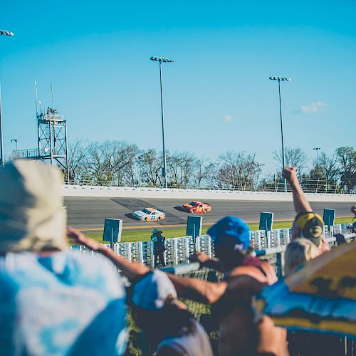 Spectators cheer at a car race on a sunny day, with three race cars speeding on the track in the background.