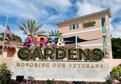 The image shows a sign reading "ART & GARDENS" with "HONORING OUR VETERANS" in front of a building with palm trees and a flag.