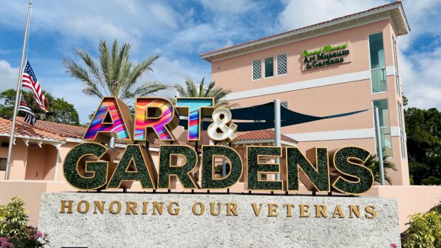 The image shows a sign reading "ART & GARDENS" with "HONORING OUR VETERANS" in front of a building with palm trees and a flag.