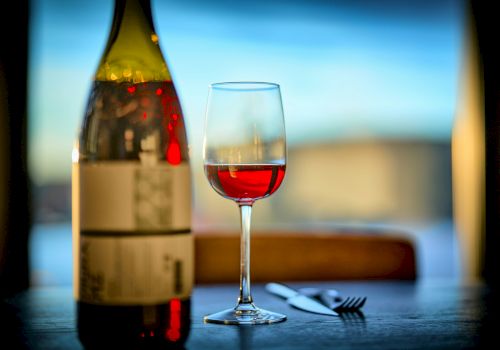 A bottle and a glass of red wine sit on a table alongside a knife and fork, with a blurred background and soft lighting.