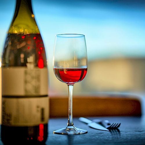 A bottle and a glass of red wine sit on a table alongside a knife and fork, with a blurred background and soft lighting.