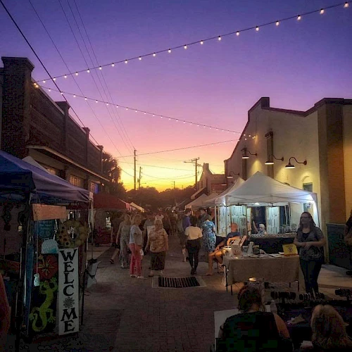 A street market at sunset with people shopping under string lights and tents, against a vibrant purple and yellow sky.