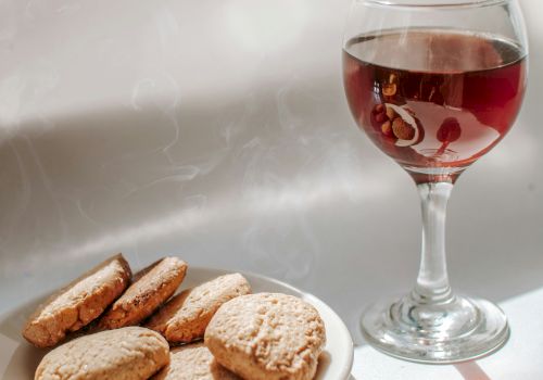 A plate of cookies and a glass of red wine are placed on a white surface, with a plant casting a shadow on the wall.