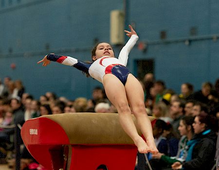 A gymnast in mid-air during a vault routine at a competition, with spectators watching intently.