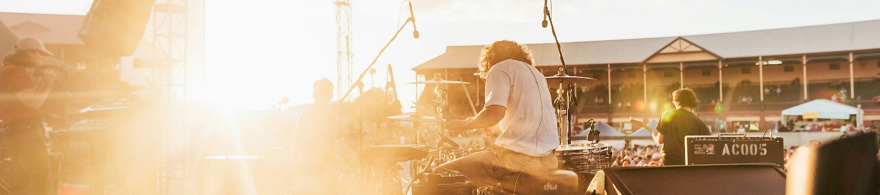 A drummer performs energetically at an outdoor stage during sunset, with equipment visible and a crowd in the background.