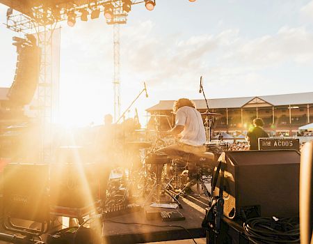 A drummer performs energetically at an outdoor stage during sunset, with equipment visible and a crowd in the background.