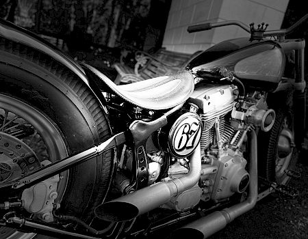 A black and white image shows a vintage motorcycle with a custom seat and the number "67" on the side.