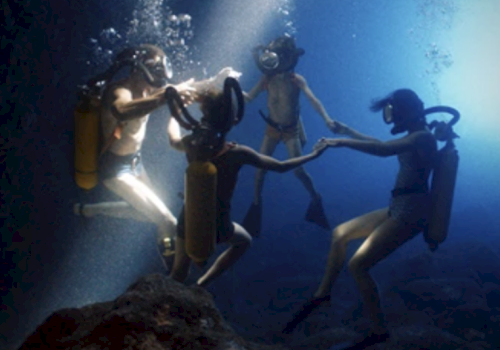 Four scuba divers are underwater, holding hands in a circle with sunlight streaming down, creating a serene and mystical scene.