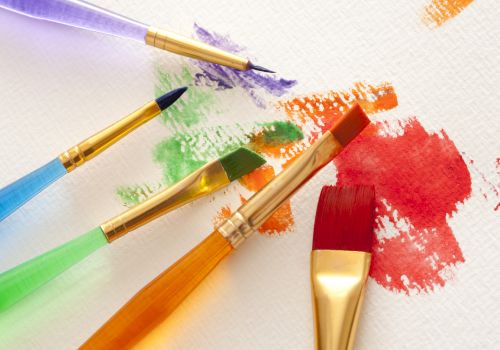 The image shows paintbrushes with colorful handles, resting on paper with red, orange, green, and purple paint streaks and splatters.