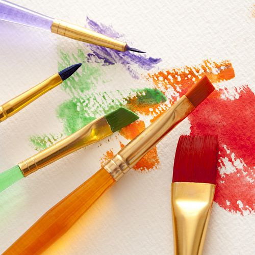 The image shows paintbrushes with colorful handles, resting on paper with red, orange, green, and purple paint streaks and splatters.