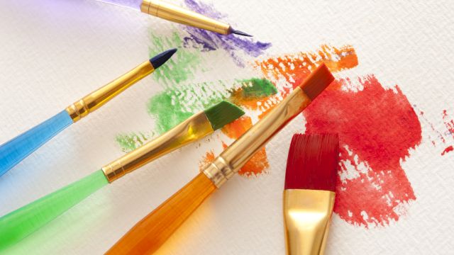 The image shows paintbrushes with colorful handles, resting on paper with red, orange, green, and purple paint streaks and splatters.