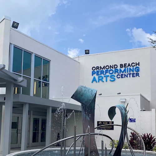 The image shows the Ormond Beach Performing Arts Center with a modern sculpture and fountain in front, under a partly cloudy sky.