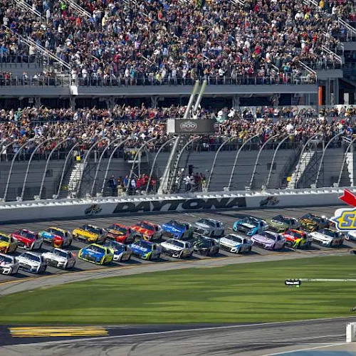 Daytona racetrack crowded with NASCAR stock cars lined up near a Sunoco logo, with packed grandstands behind them.