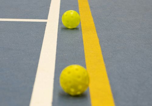 The image shows two yellow perforated balls on a blue floor with yellow and white court lines.