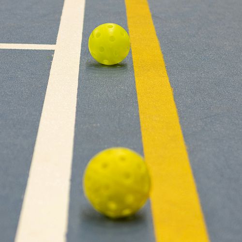 The image shows two yellow perforated balls on a blue floor with yellow and white court lines.