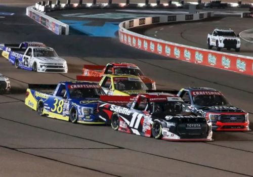 A fleet of NASCAR-style pickup trucks races on a speedway track under stadium lights.