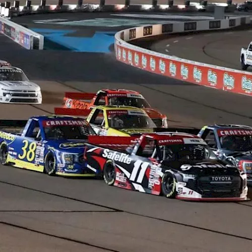 A fleet of NASCAR-style pickup trucks races on a speedway track under stadium lights.