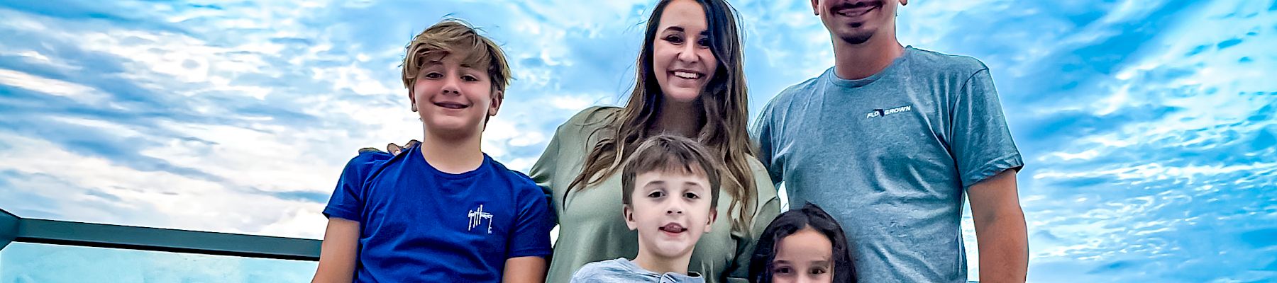 A family of five poses on a balcony overlooking the ocean, with a beautiful blue sky.