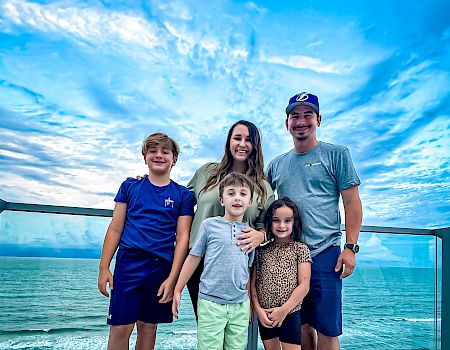 A family of five poses on a balcony overlooking the ocean, with a beautiful blue sky.