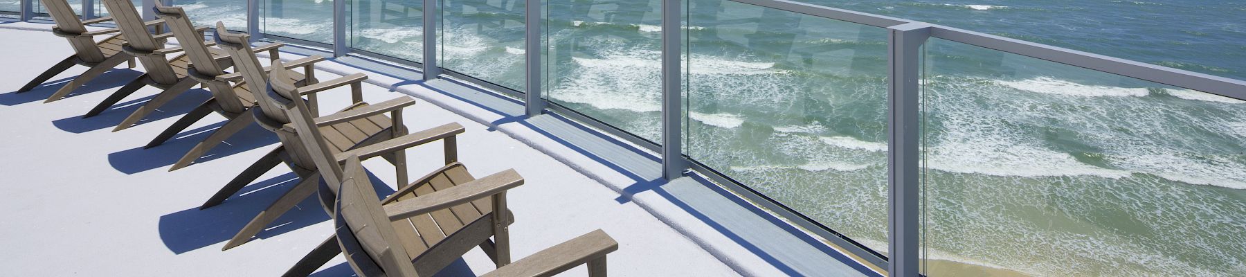 Chairs on a deck overlook an expansive ocean view with clear skies and waves hitting the shore, bordered by a glass railing.