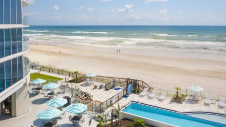 A beachfront scene with a pool, lounge chairs, umbrellas, and a sandy beach meeting the ocean.