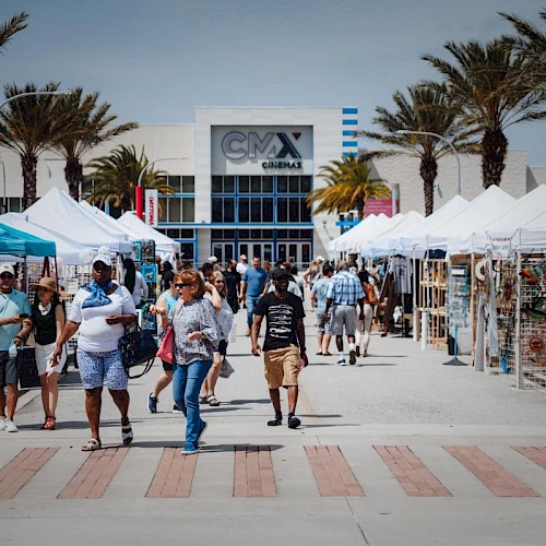 People walk through an outdoor market with tents and stalls on both sides, near a building marked "CMX Cinemas."