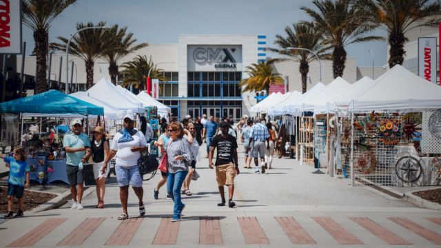 People walk through an outdoor market with tents and stalls on both sides, near a building marked "CMX Cinemas."