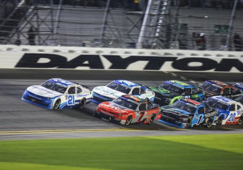 A NASCAR-style stock car race at Daytona, with several cars packed together on the track at high speed.