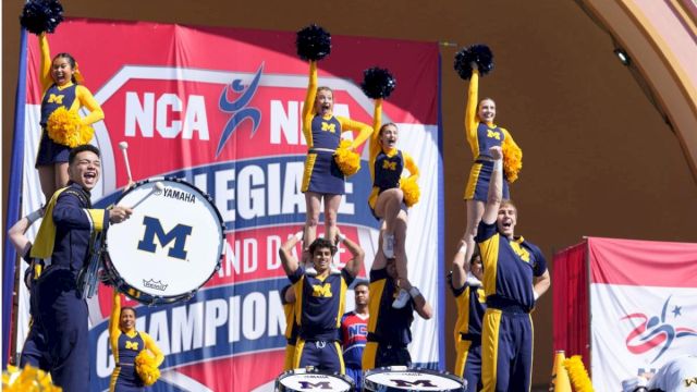 Cheerleaders and a drumline perform at the NCA & NDA Collegiate Championships in front of a banner and an excited crowd.