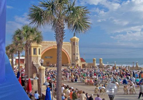 A crowded seaside walkway with palm trees and a big arched building, with people gathered along the beach under blue sky.