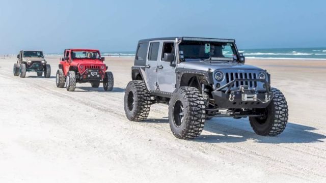 Three off-road vehicles drive on a sandy beach, with ocean waves visible in the background, under a clear blue sky.