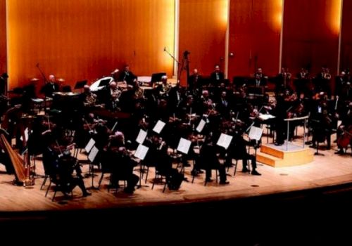 A large orchestra of musicians with strings and brass performs on a concert hall stage under warm lights.