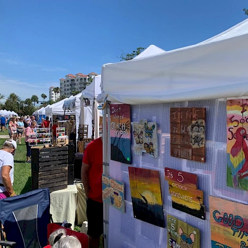 An outdoor art market with people browsing under tents, displaying colorful paintings and crafts for sale in a sunny setting.
