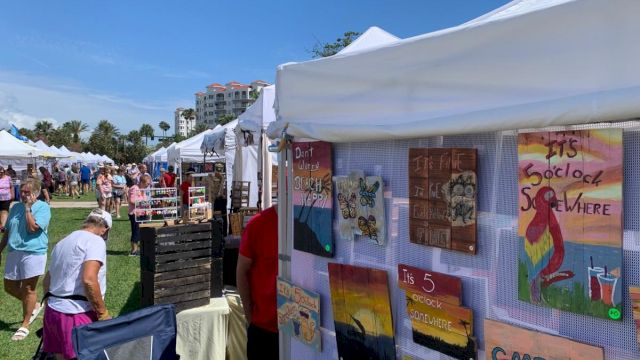 An outdoor art market with people browsing under tents, displaying colorful paintings and crafts for sale in a sunny setting.