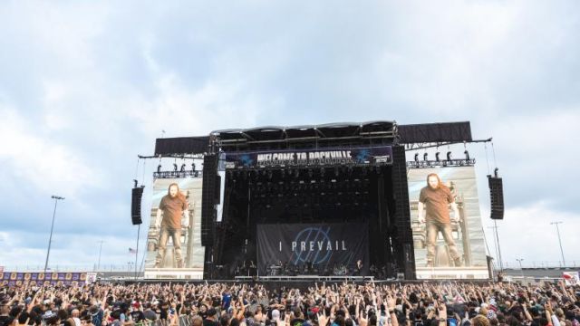 A large outdoor concert with a crowd in front of a stage displaying screens and a backdrop reading "I Prevail".