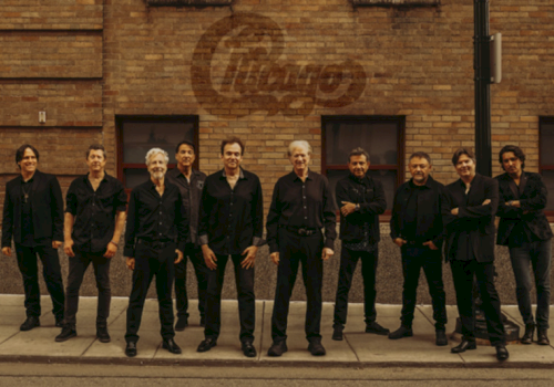 A group of eight men pose on a sidewalk in front of a brick building with a faded logo painted on the wall.