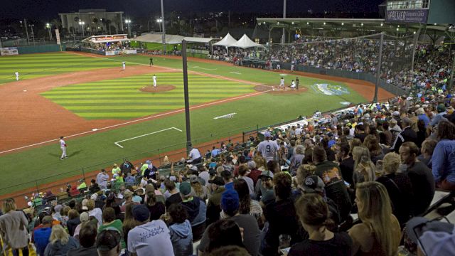 A nighttime baseball game is underway with a packed crowd watching from the stands and players on the field.