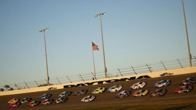 Race cars appear on a track, with the American flag and "Daytona" visible, under clear skies and floodlights.