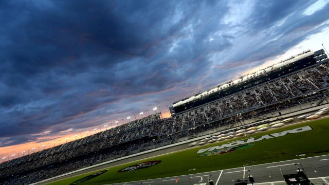 A racetrack under a dramatic, cloudy sky with a grandstand full of spectators, and parts of the track are illuminated by stadium lights.