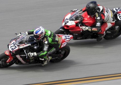 Two motorcyclists are racing on a track, one in green and black gear, the other in red and white, leaning into a curve.
